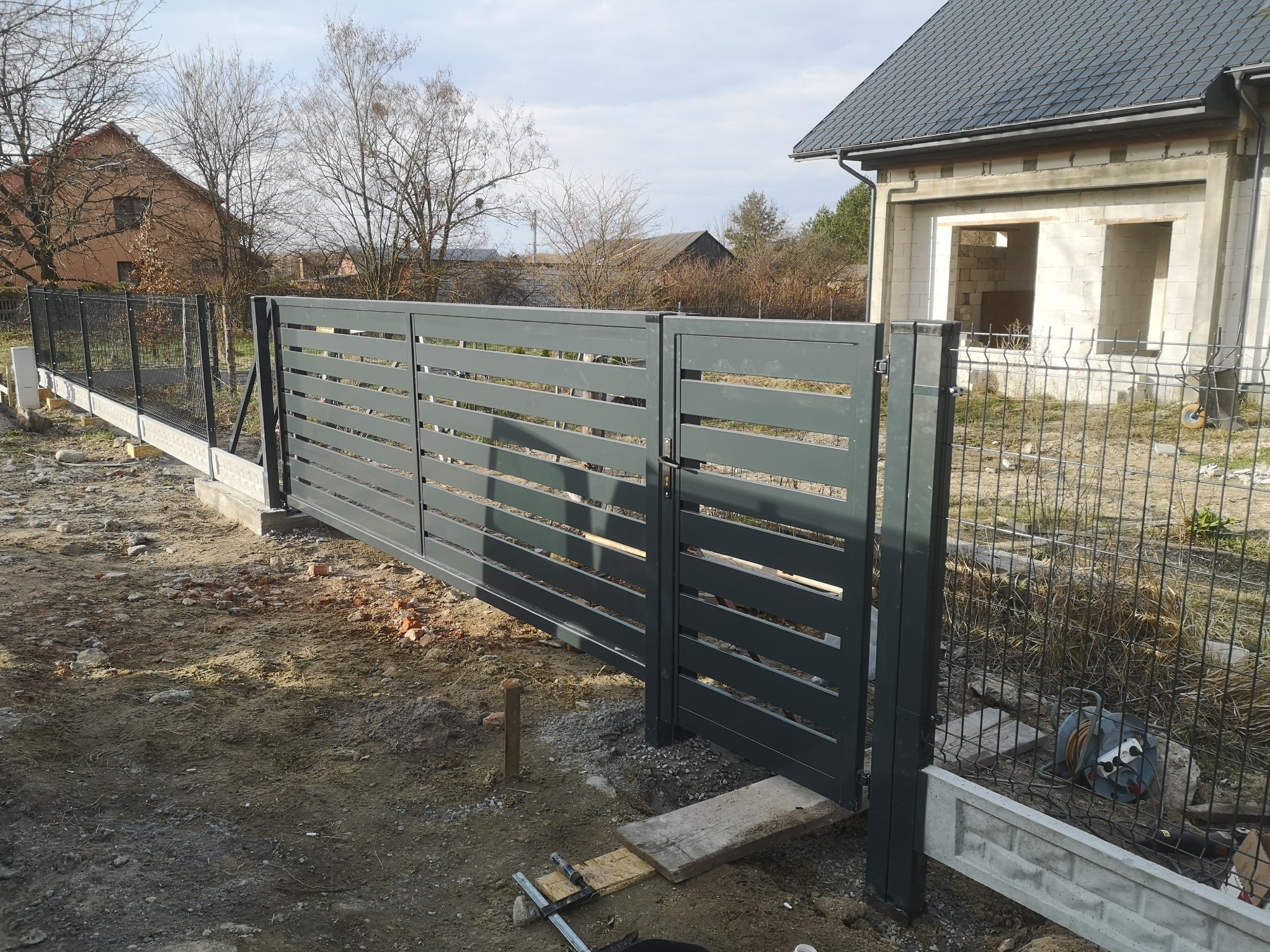 Nowoczesna, przesuwna brama w kolorze grafitowym z poziomymi szczeblami, widoczna na tle surowego, nieotynkowanego domu w trakcie budowy i częściowo postawionego ogrodzenia z siatki.