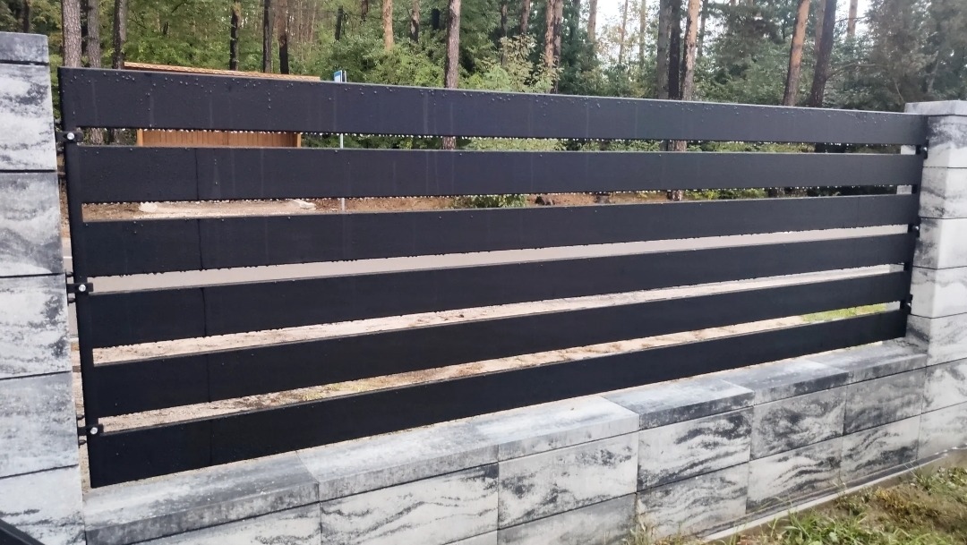 Nowoczesne ogrodzenie panelowe w kolorze antracytowym z betonowymi słupkami i podmurówką z szarej kostki imitującej marmur, na tle lasu.
