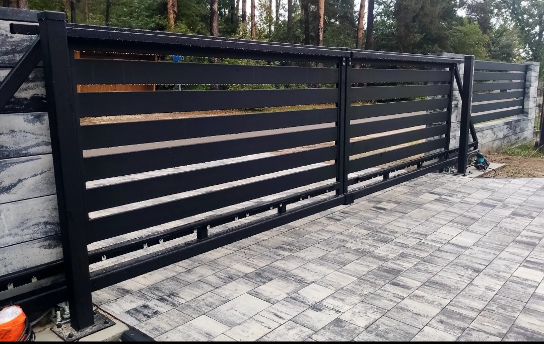 Nowoczesna, przesuwna brama wjazdowa z poziomych, czarnych profili, zamontowana na posesji z kostką brukową i ogrodzeniem z betonowych bloczków.