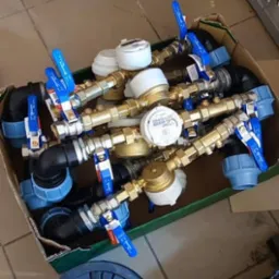 Karton pełen złączek, zaworów i liczników wody z niebieskimi uchwytami, obok leży dekiel od studzienki kanalizacyjnej i skrzynka narzędziowa.