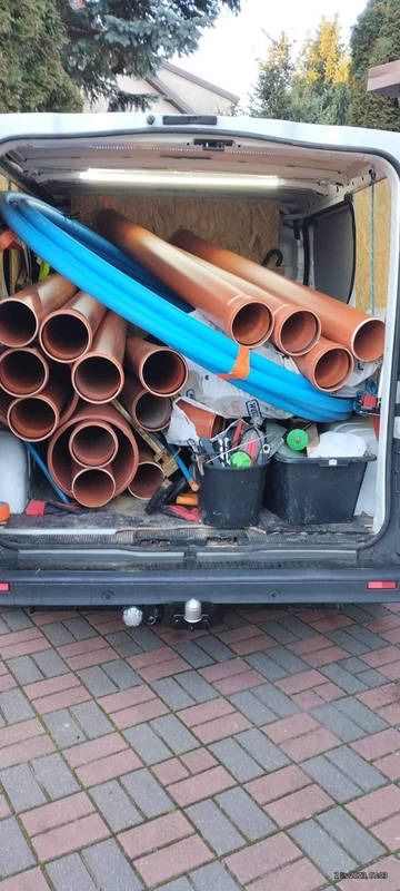 Wnętrze furgonetki wypełnionej brązowymi rurami kanalizacyjnymi i niebieskimi rurami wodnymi, widok z tyłu pojazdu na kostkę brukową.