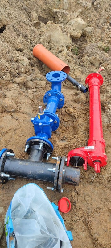 Elementy instalacji wodociągowej: niebieskie i czerwone rury z kołnierzami, pomarańczowa rura oraz czarne złączki leżące na ziemi obok wykopu.