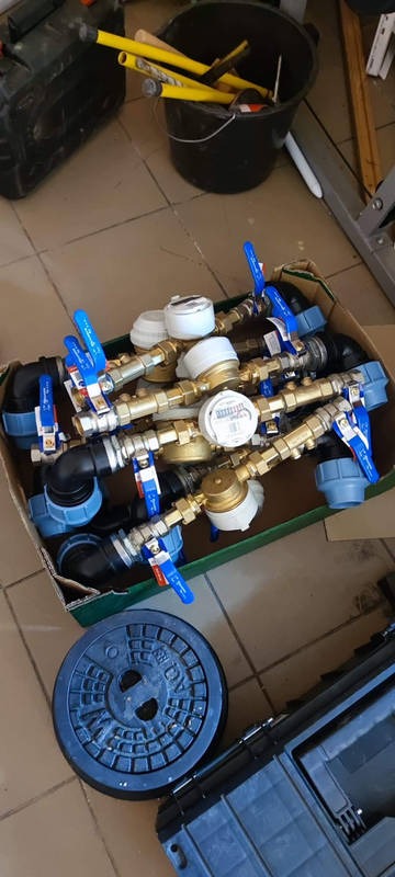 Karton pełen złączek, zaworów i liczników wody z niebieskimi uchwytami, obok leży dekiel od studzienki kanalizacyjnej i skrzynka narzędziowa.