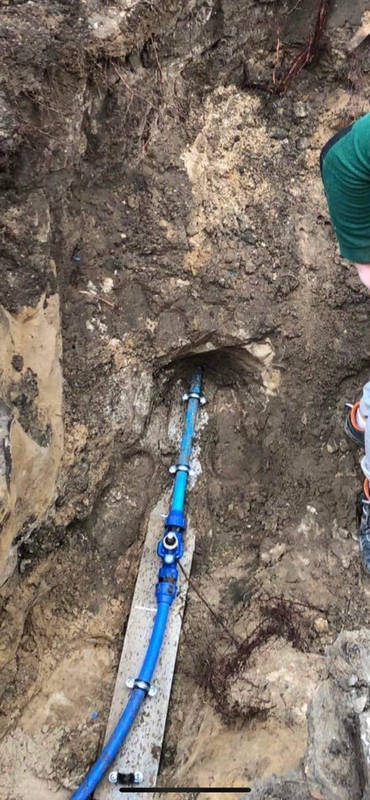 Niebieska rura wodociągowa z zaworem, zamocowana klamrami do deski, wystająca z wykopu w ziemi. Widoczne korzenie i fragment sylwetki osoby.