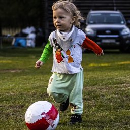 Piesik - Mały chłopiec z blond włosami biegnie po trawie w kierunku piłki nożnej z napisem 'Polska', w tle zaparkowany ciemny samochód.