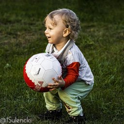 Piesik - Uśmiechnięty chłopiec z kręconymi blond włosami trzyma dużą biało-czerwoną piłkę na tle zielonej trawy.