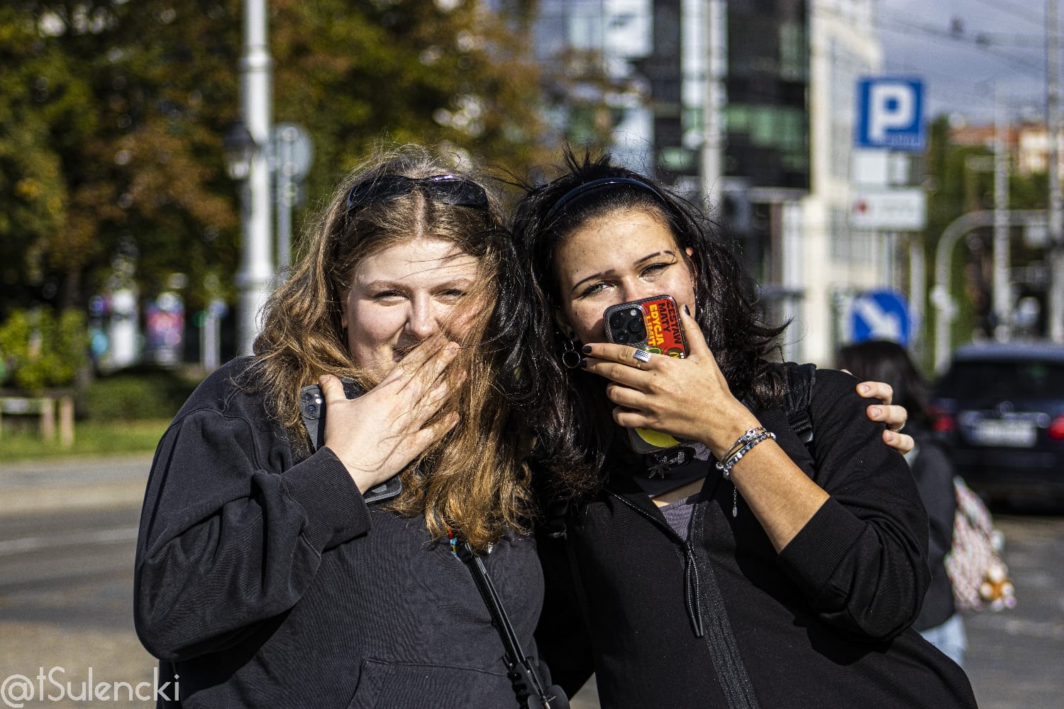 Dwie uśmiechnięte kobiety w czarnych bluzach, stojące na tle miejskiej ulicy, zasłaniają twarze dłońmi i telefonem w żółtym etui.