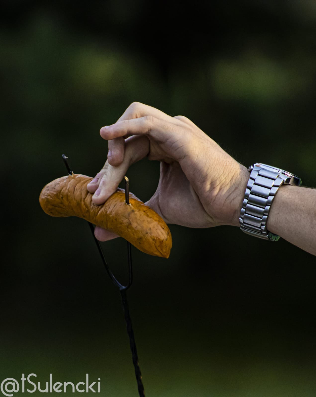 Ręka w zegarku trzymająca przypieczoną kiełbasę nabitą na widelec do grilla na tle ciemnej zieleni.