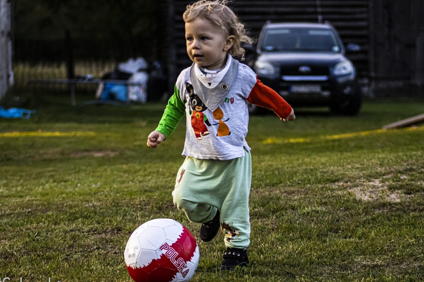 Mały chłopiec z blond włosami biegnie po trawie w kierunku piłki nożnej z napisem 'Polska', w tle zaparkowany ciemny samochód.