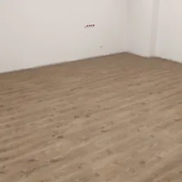 Jasnobrązowe panele podłogowe ułożone w pustym pokoju z białymi ścianami, widoczne gniazdka elektryczne i listwy przypodłogowe.