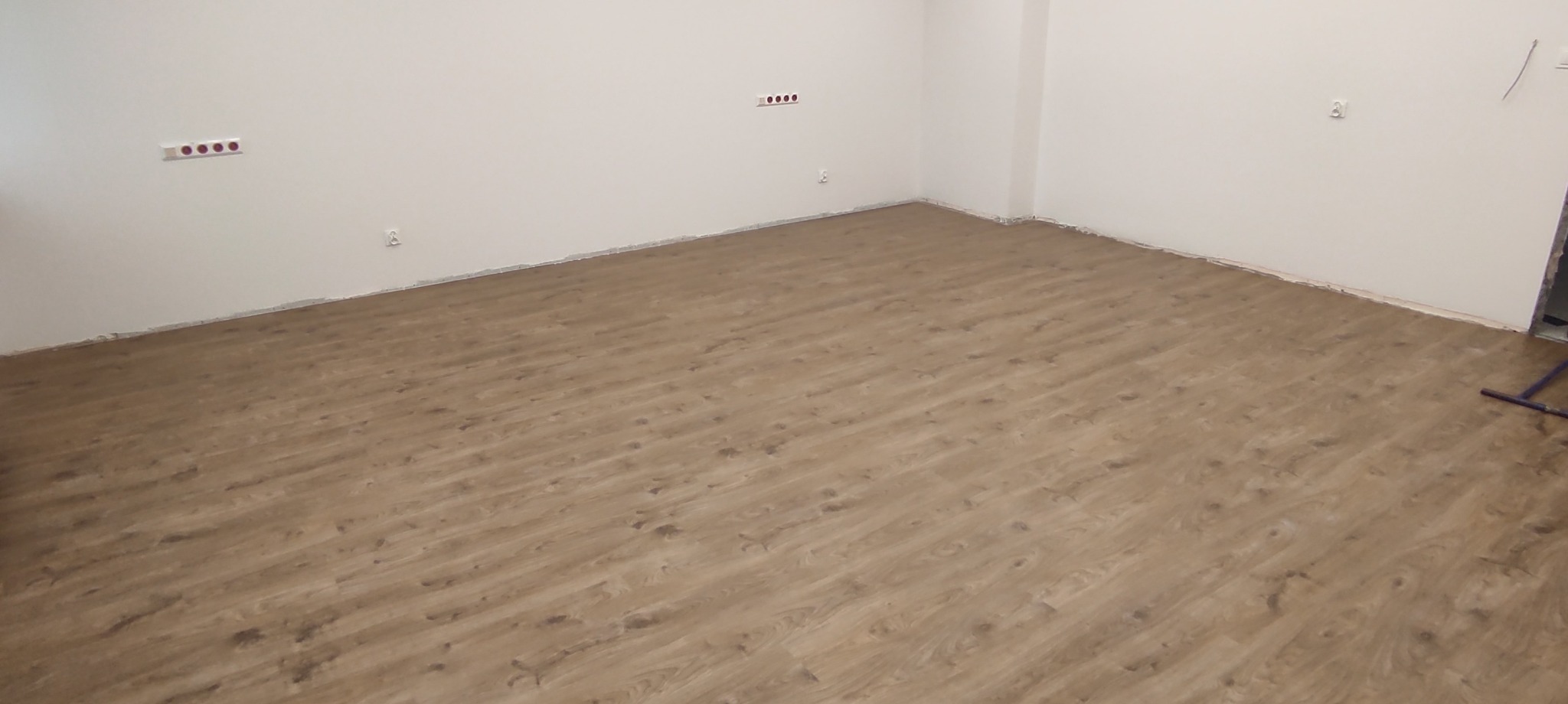 Jasnobrązowe panele podłogowe ułożone w pustym pokoju z białymi ścianami, widoczne gniazdka elektryczne i listwy przypodłogowe.