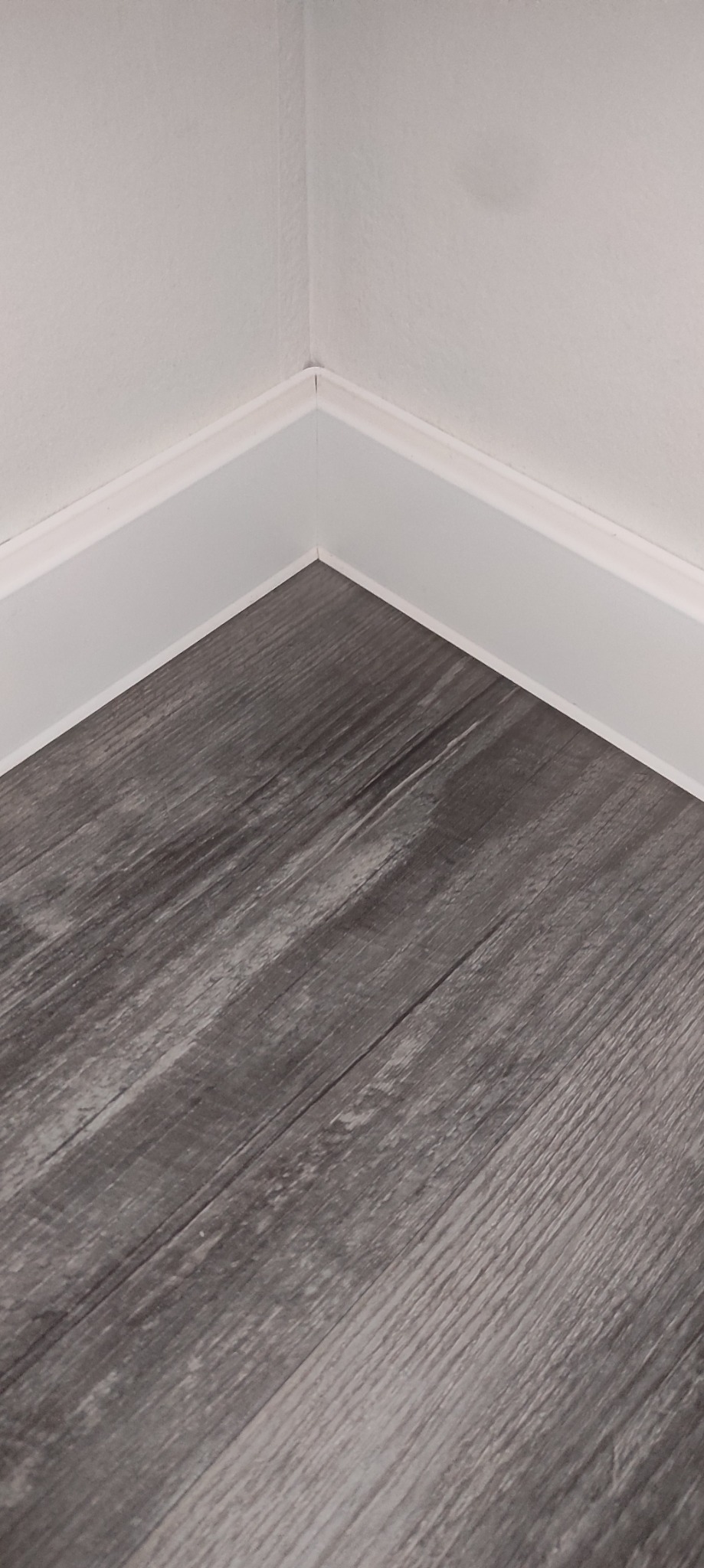 Szare panele podłogowe imitujące drewno, ułożone przy białej listwie przypodłogowej w rogu pomieszczenia.
