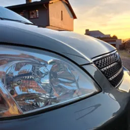 Błyszczący reflektor odnowionego, szarego samochodu na tle domu i zachodzącego słońca.