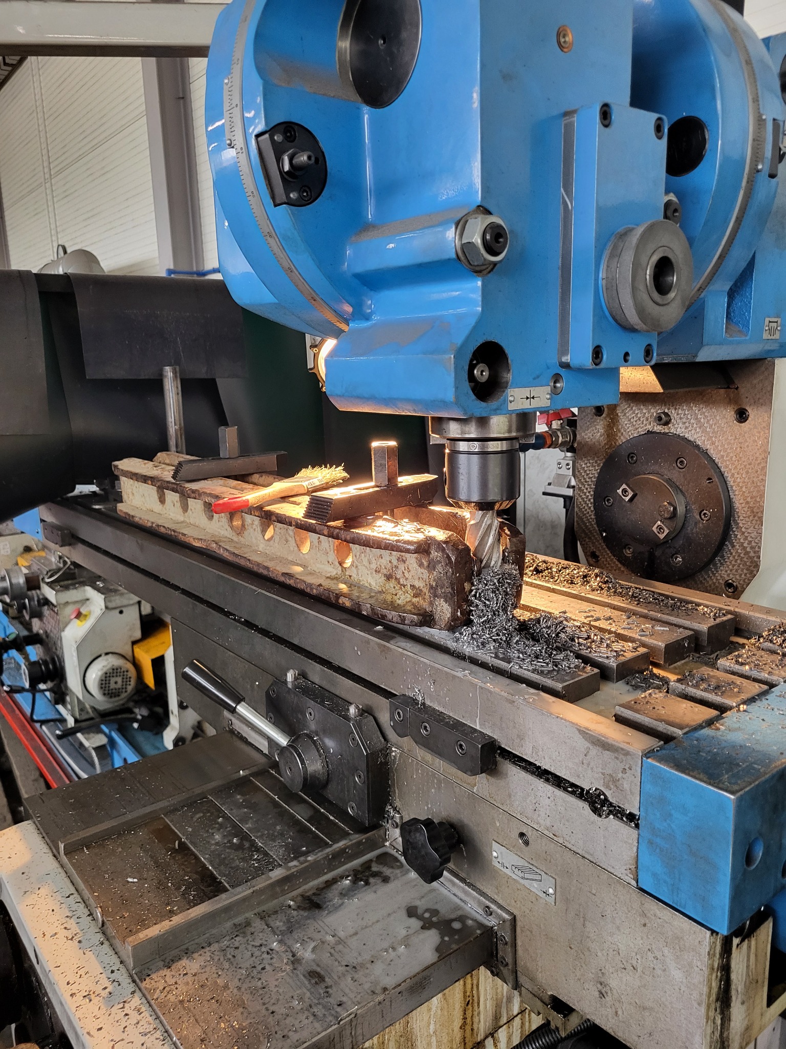 Frezarka CNC w trakcie obróbki metalowego elementu, wióry na stole roboczym, widoczna szczotka do czyszczenia.