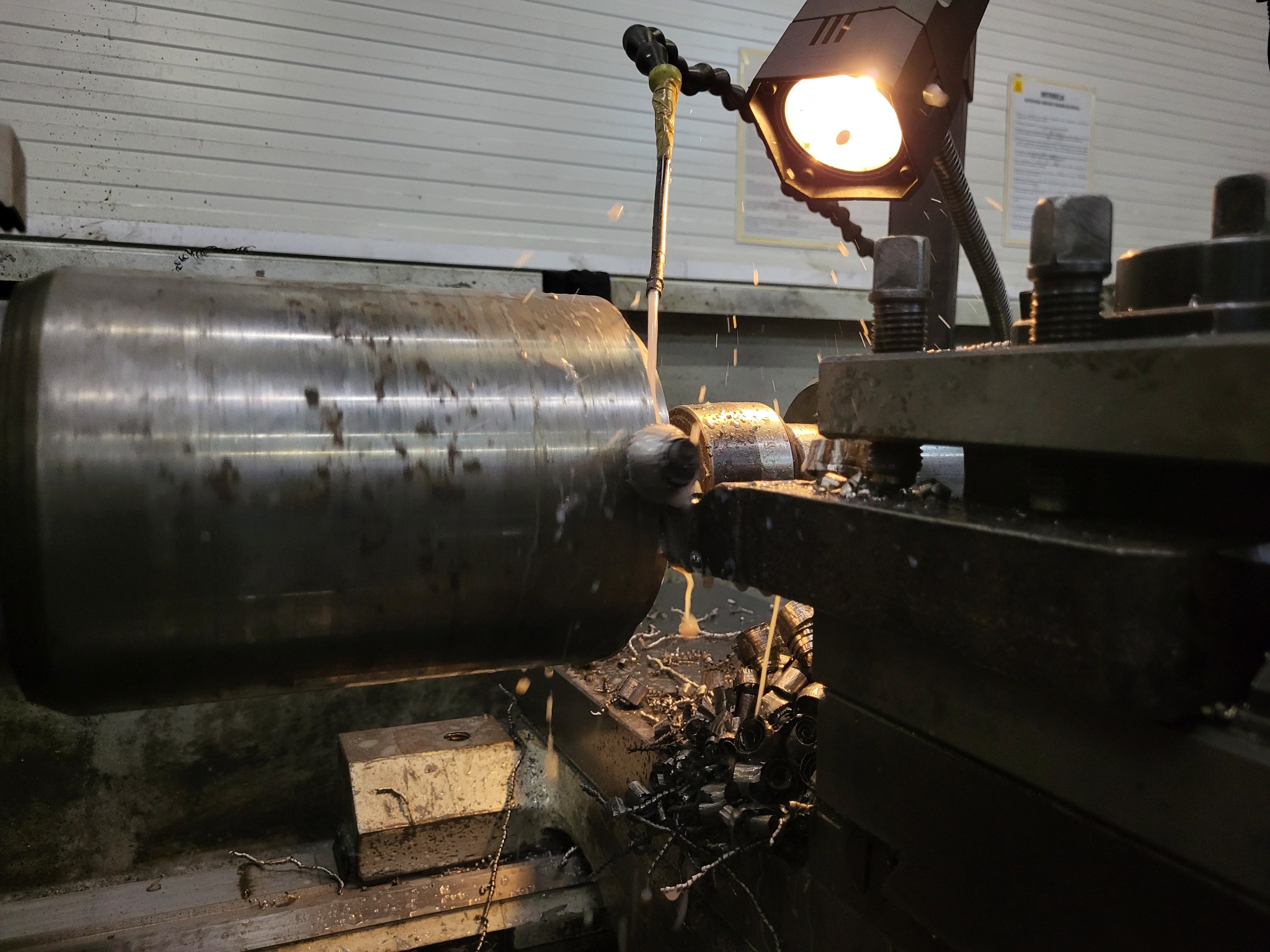 Obróbka metalu na tokarce: cylinder obracający się w uchwycie, strumień chłodziwa i opiłki metalu w obszarze roboczym, oświetlone lampą warsztatową.