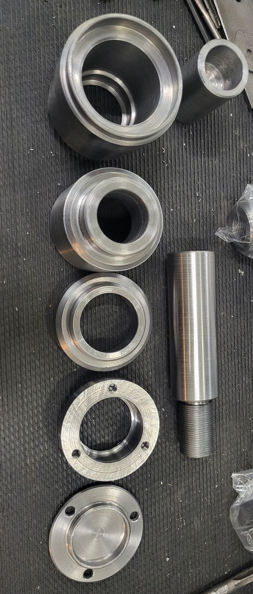 Metalowe pierścienie i tuleja z gwintem, ułożone na ryflowanej powierzchni, widoczne ślady obróbki skrawaniem.