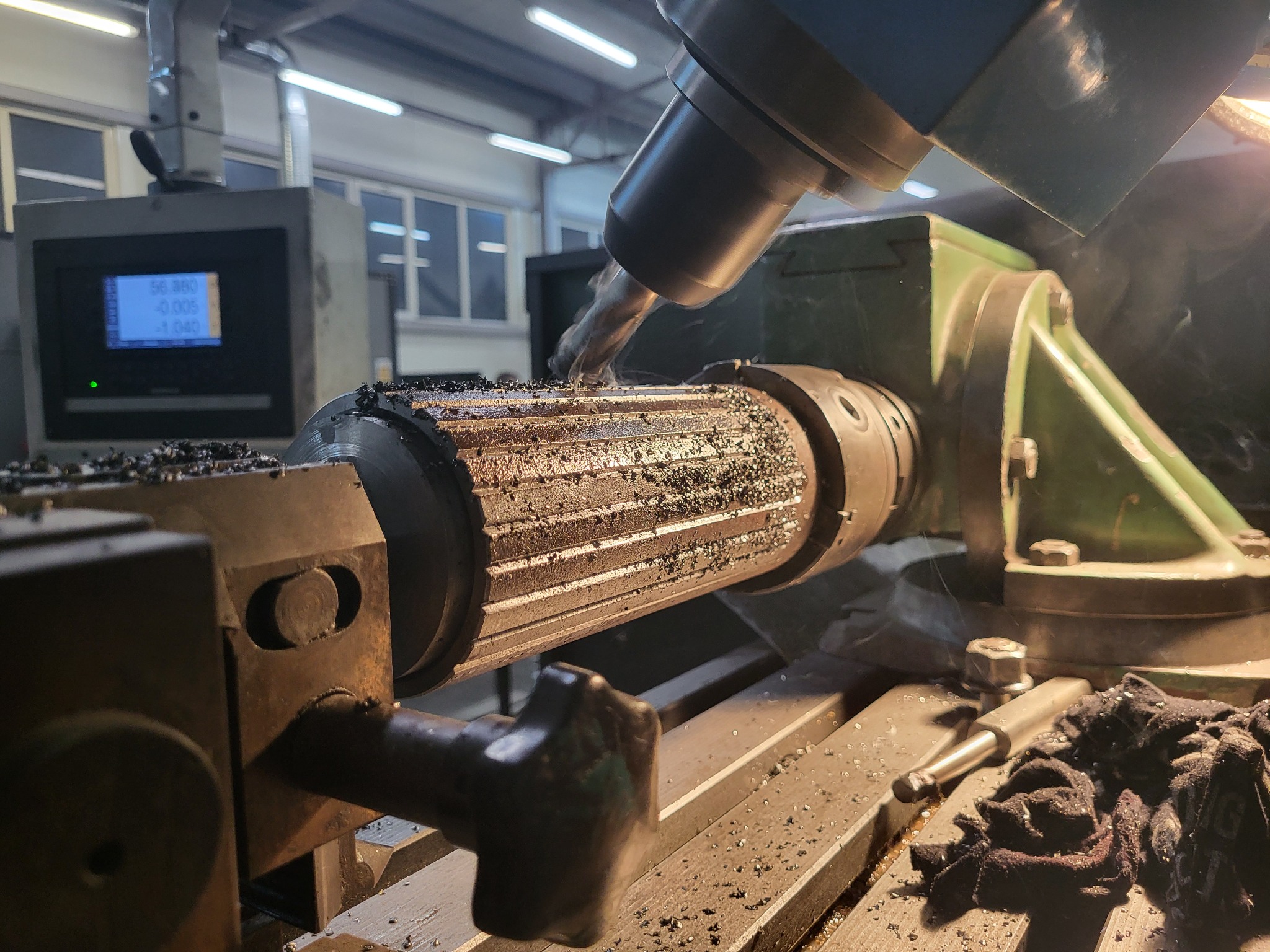 Frezarka CNC w trakcie obróbki metalowego walca, widoczne opiłki, chłodziwo i panel sterowania maszyny.