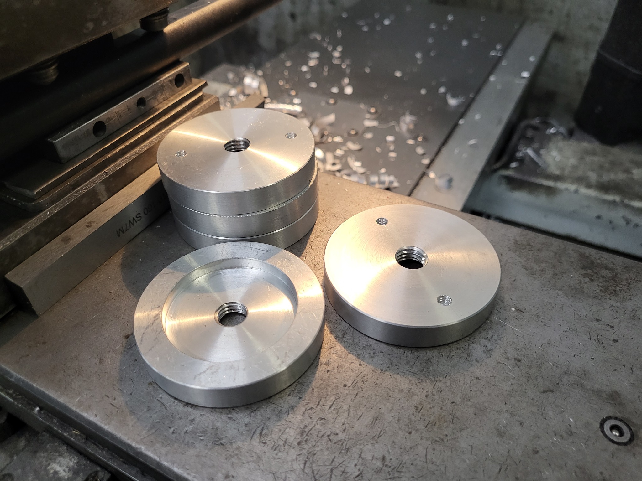 Aluminiowe, okrągłe elementy z gwintowanymi otworami, ułożone na metalowym stole warsztatowym, w tle wióry i narzędzia.
