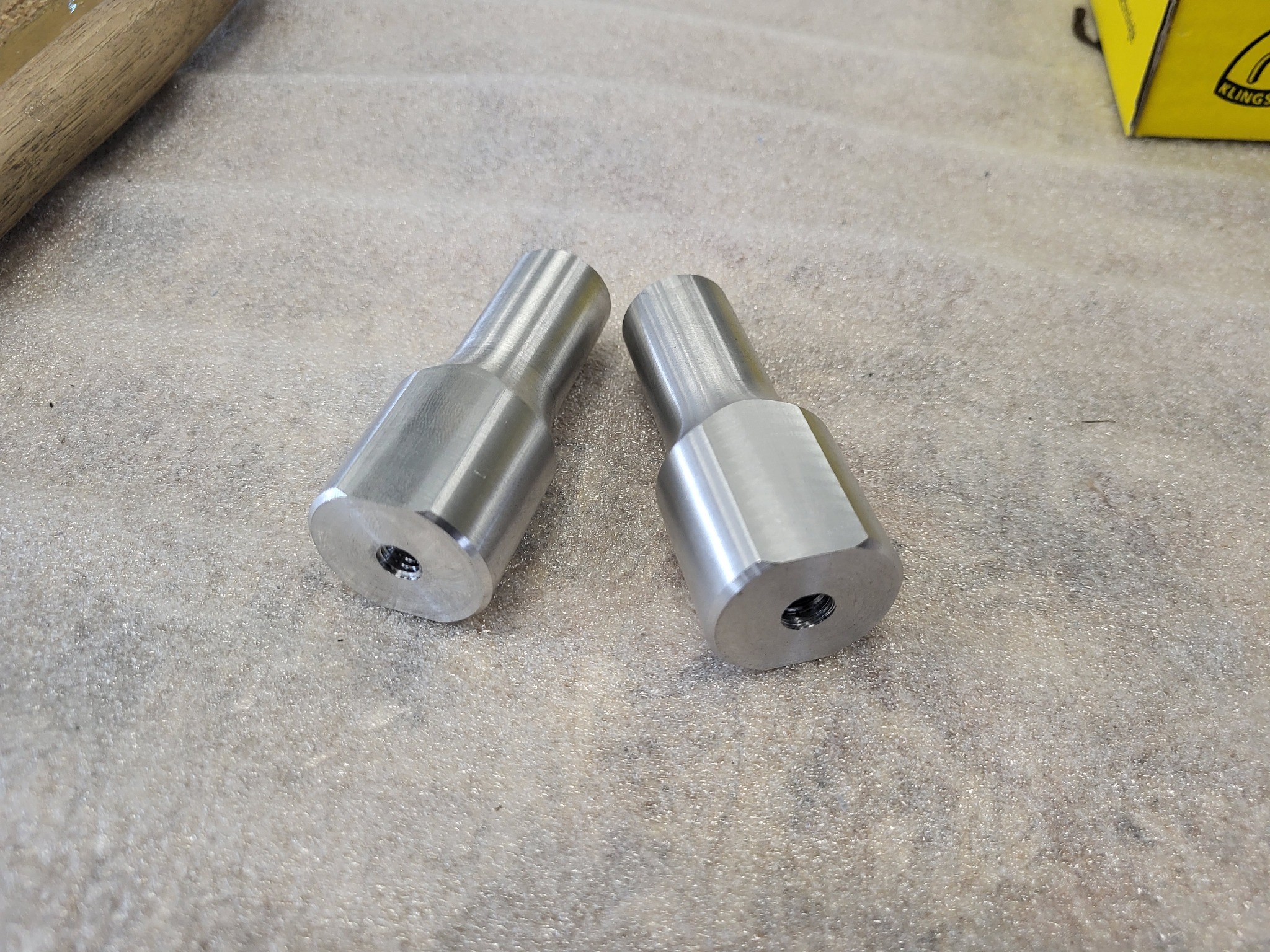 Dwa identyczne, metalowe elementy z gwintowanymi otworami na szarym, teksturowanym tle, z widocznym fragmentem drewnianego narzędzia i żółtego pudełka w tle.