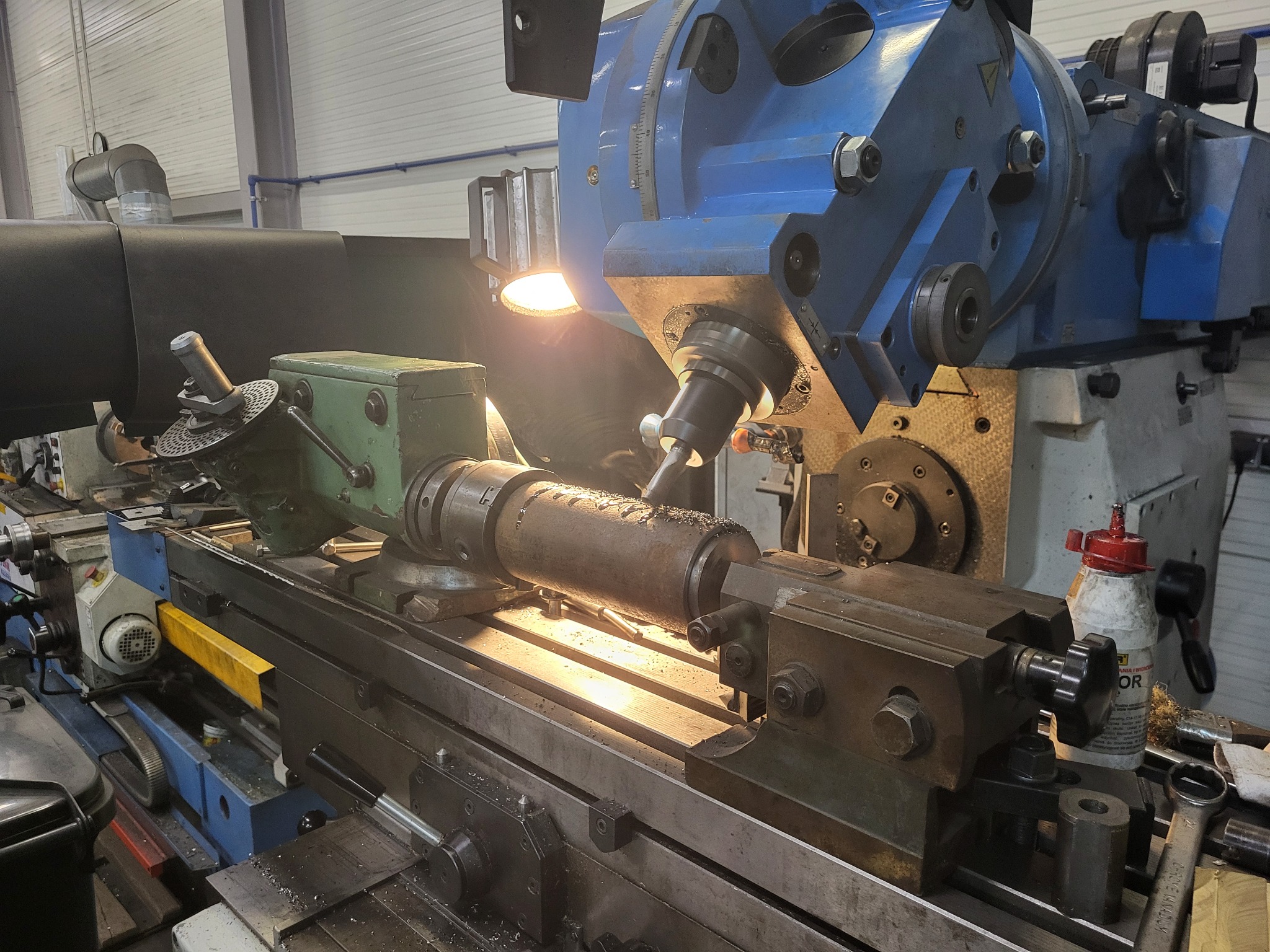 Obrabiany metalowy wałek w uchwycie tokarki z widocznymi wiórami, oświetlony lampą, z narzędziami i akcesoriami w tle.