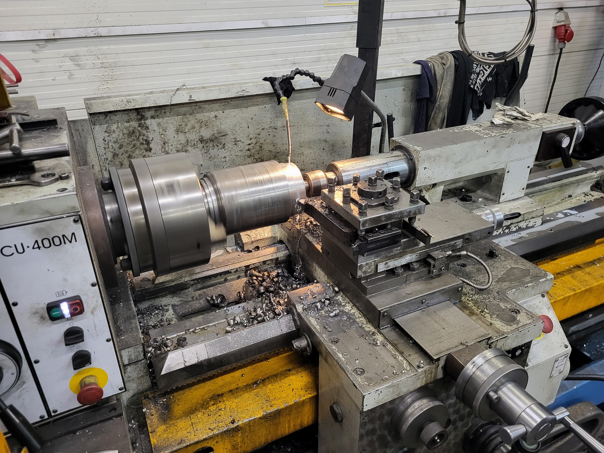 Metalowy element obrabiany na tokarce CU-400M pod oświetleniem lampki, wióry na stole roboczym.