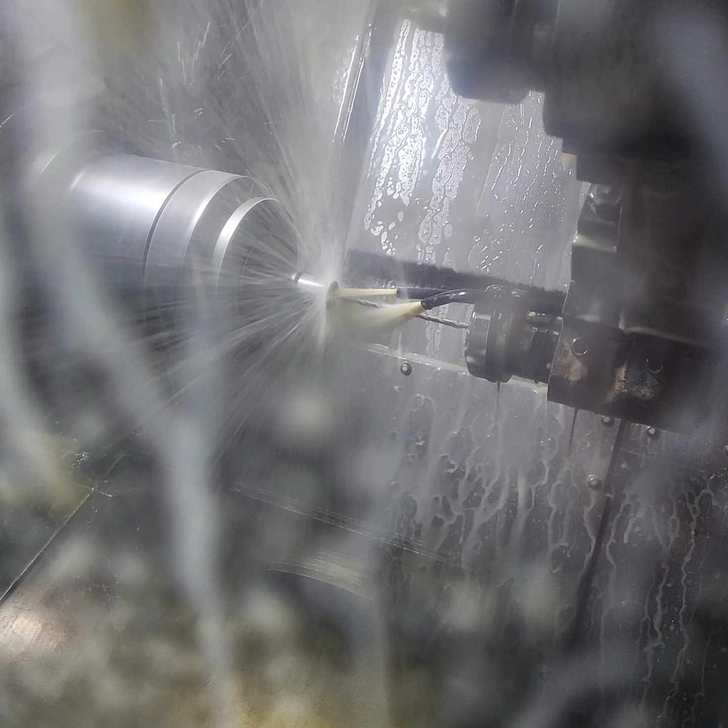 Metalowy element obrabiany w tokarce CNC z użyciem chłodziwa, widoczne wióry i oprysk cieczy, oświetlenie halogenowe.