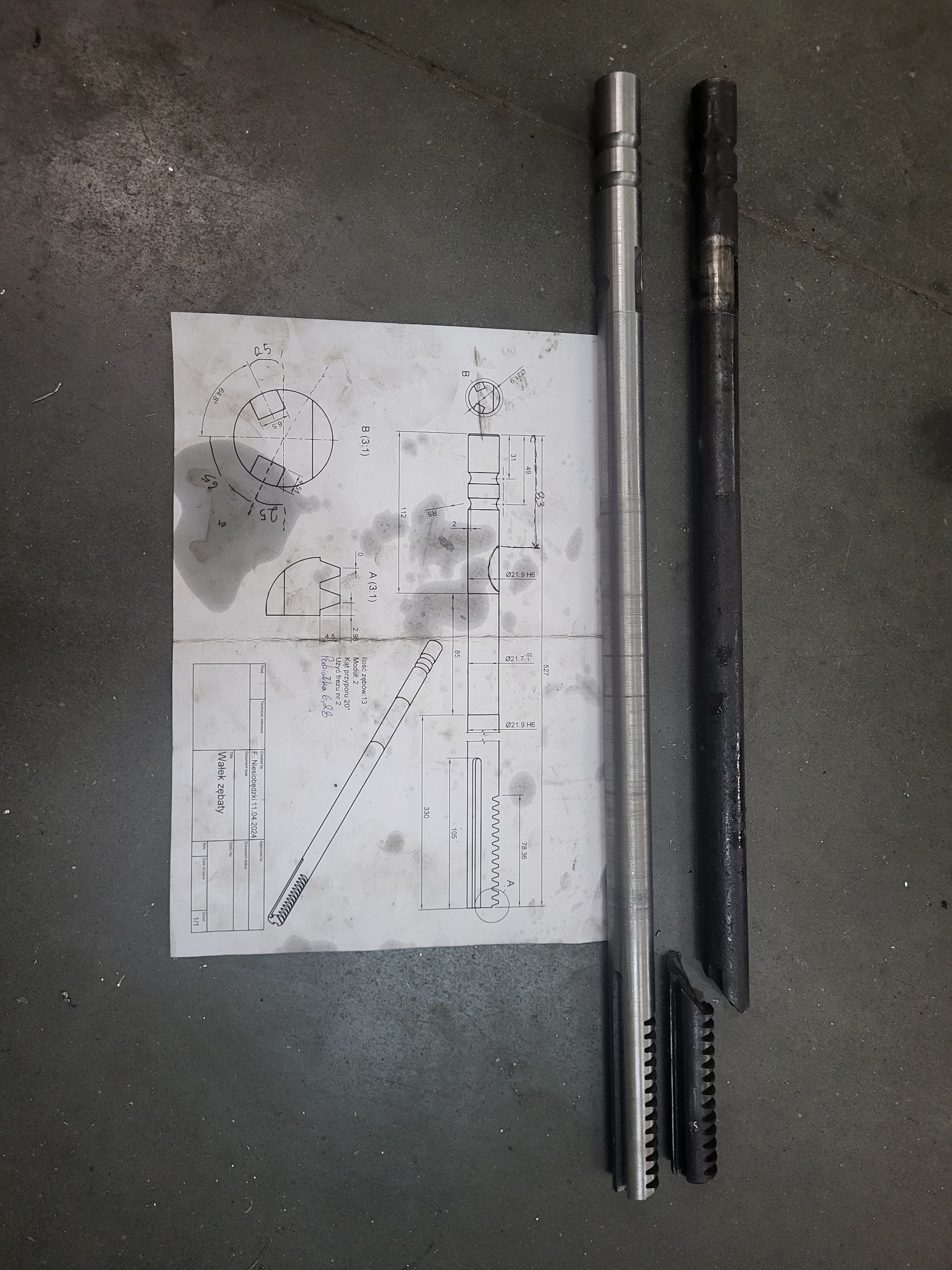 Wałek zębaty po obróbce, leżący obok rysunku technicznnego z naniesionymi wymiarami, obok drugi wałek po obróbce wstępnej.