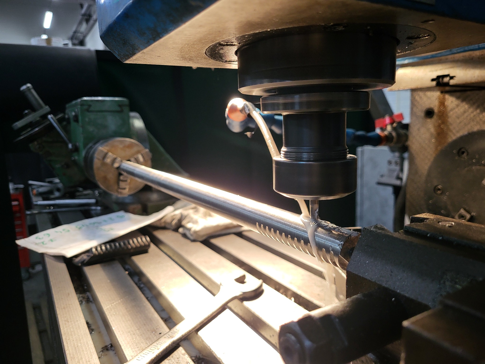 Frezowanie CNC metalowego wałka z zębatką, chłodziwo spływa po obrabianym elemencie, widoczny klucz płaski i notatki na stole frezarki.