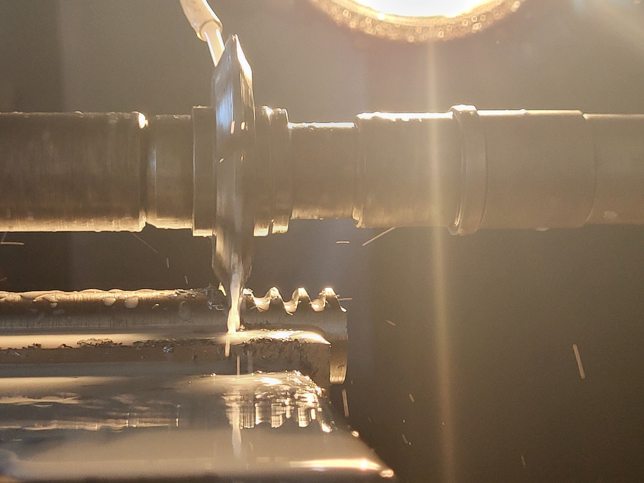 Metalowy wał obrabiany przez ostrze z chłodziwem, widoczne iskry i odbicia w cieczy, oświetlenie halogenowe w tle.