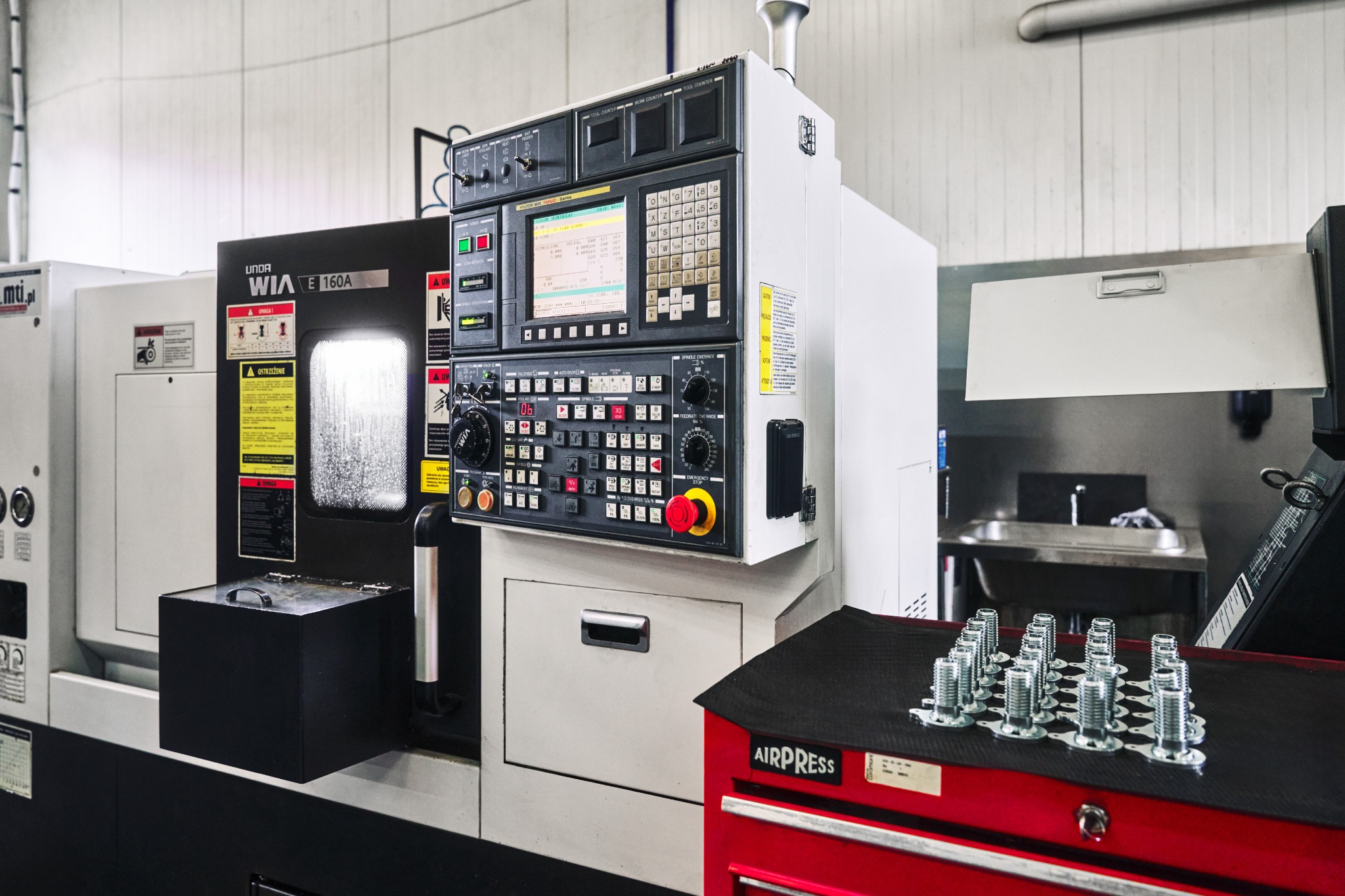 Precyzyjna obrabiarka CNC firmy UNDA WIA E160A z panelem sterowania, obok na czerwonym wózku warsztatowym ułożone w rzędach metalowe śruby z gwintem.