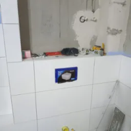 Ściana w trakcie układania płytek w łazience, widoczne narzędzia, puszka pod stelaż WC Geberit oraz wyprowadzenia rur kanalizacyjnych.