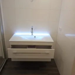 Biała umywalka z oświetleniem LED i otwartą szufladą w nowoczesnej łazience z białymi, kwadratowymi płytkami ściennymi i ciemną podłogą imitującą drewno.