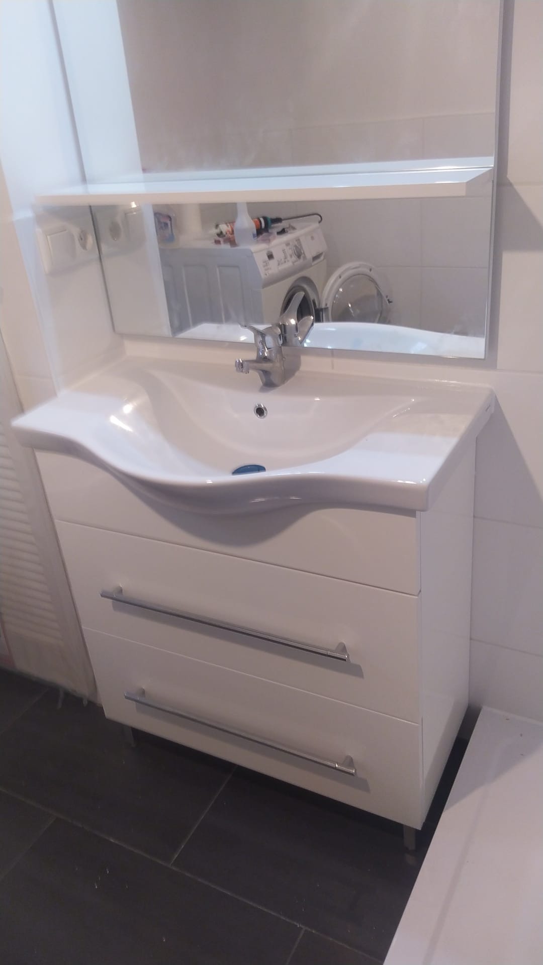Biała szafka umywalkowa z trzema szufladami i lustrem, w którym odbija się pralka w łazience z ciemną podłogą.