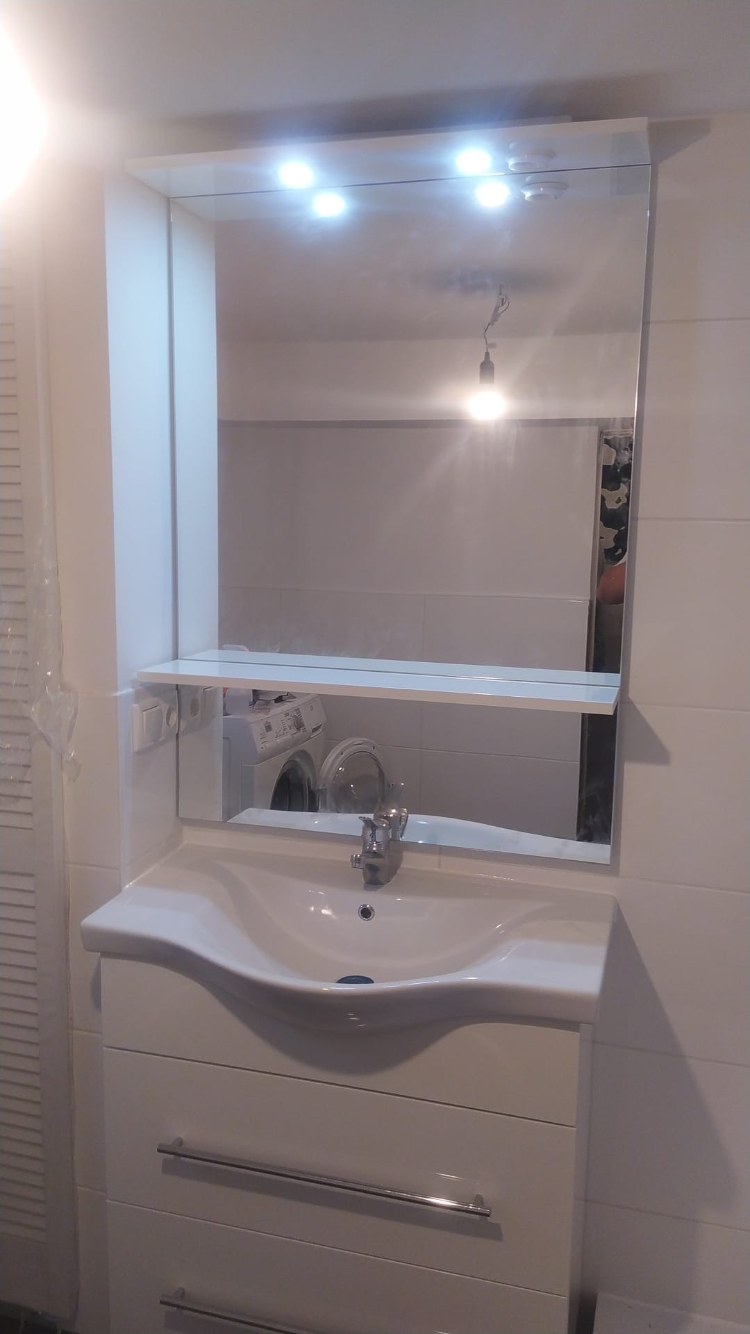 Biała umywalka z szafką i lustrem z oświetleniem w łazience, w lustrze odbija się pralka z otwartymi drzwiczkami.