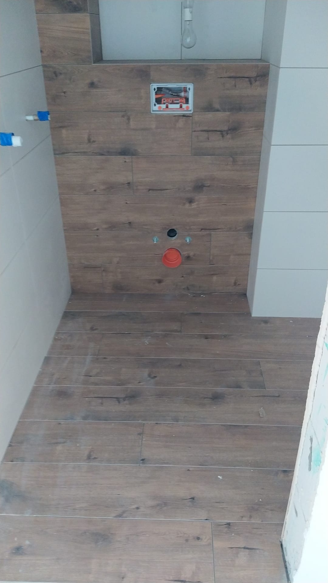 Ściana i podłoga w trakcie układania płytek imitujących drewno, widoczne instalacje sanitarne i elektryczne, surowa żarówka zwisająca z sufitu.