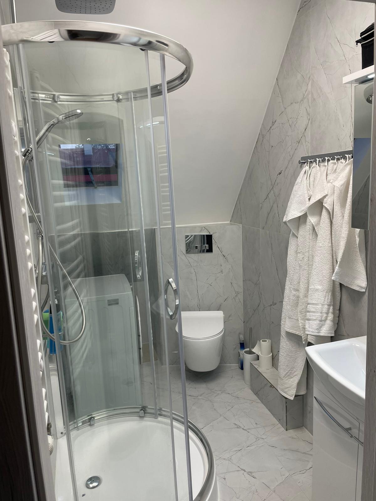 Nowoczesna łazienka na poddaszu z kabiną prysznicową, toaletą podwieszaną i marmurowymi płytkami na ścianach, pralka widoczna w kabinie.