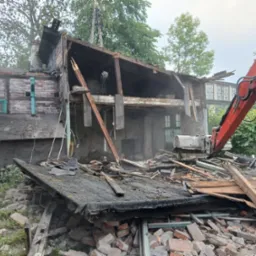 Częściowa rozbiórka budynku z cegły, widoczny gruz i elementy konstrukcyjne. Czerwony wysięgnik koparki Kubota w trakcie pracy. Fragmenty ścian z oknami.