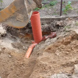 Wykop z pomarańczowymi rurami kanalizacyjnymi, widoczna łyżka koparki. Prace ziemne w toku, teren z piaskiem i gruzem, siatka ogrodzeniowa w tle.