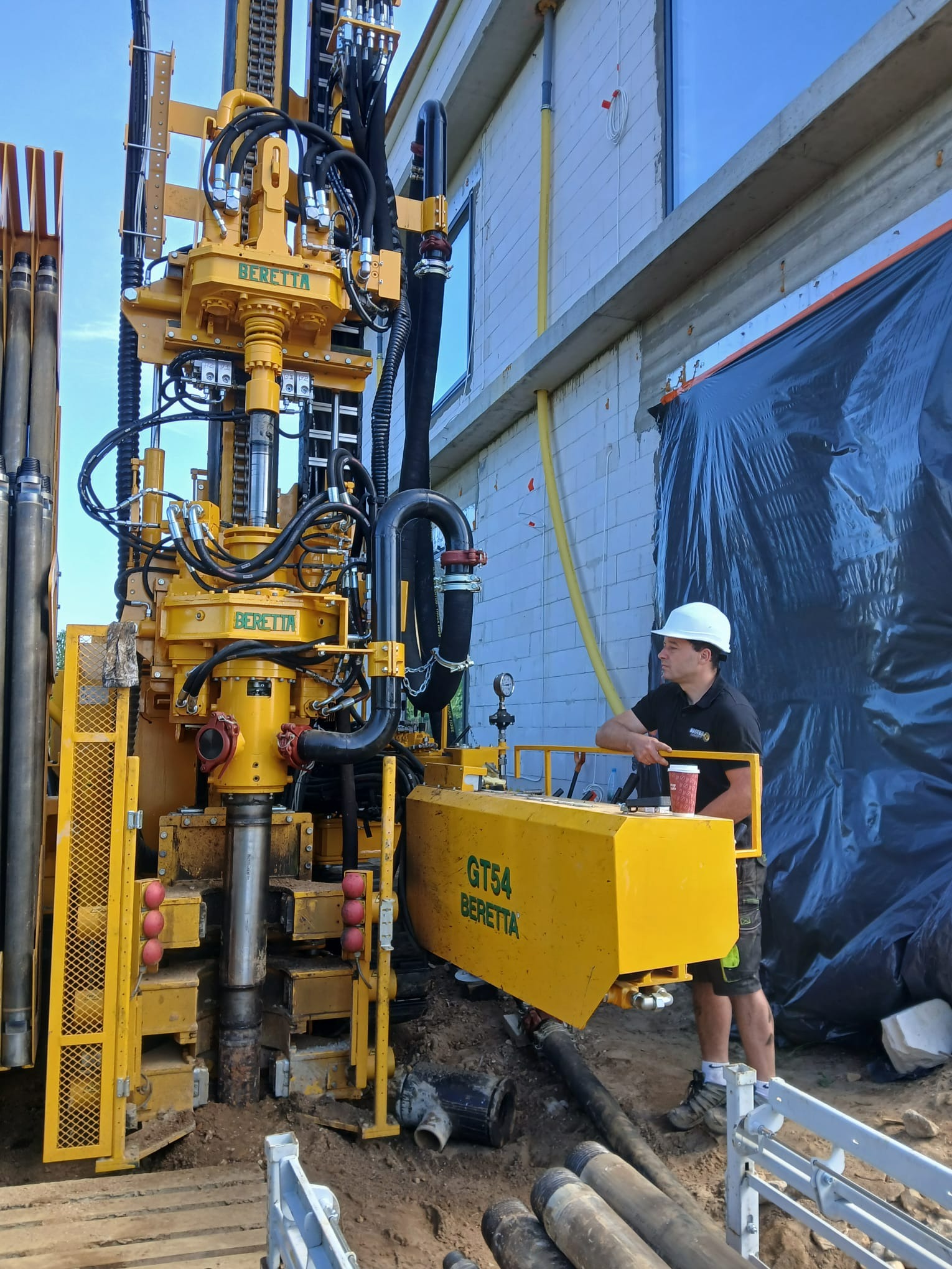 Żółta wiertnica Beretta GT54 w Oświęcimiu, obsługiwana przez operatora w kasku. W tle budynek w trakcie budowy, rury wiertnicze i czarne zabezpieczenie.