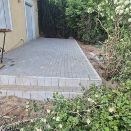 Mateusz - Jasnoszara kostka brukowa ułożona na tarasie przy domu, widoczna zagęszczarka i roślinność. Krawędzie tarasu wykończone kostką. Częściowo widoczny budynek.