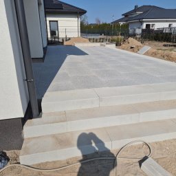 Mateusz - Jasnoszara, ułożona kostka granitowa tworzy taras z trzema schodami. W tle dom z solarnymi panelami i przygotowany teren pod nasadzenia.