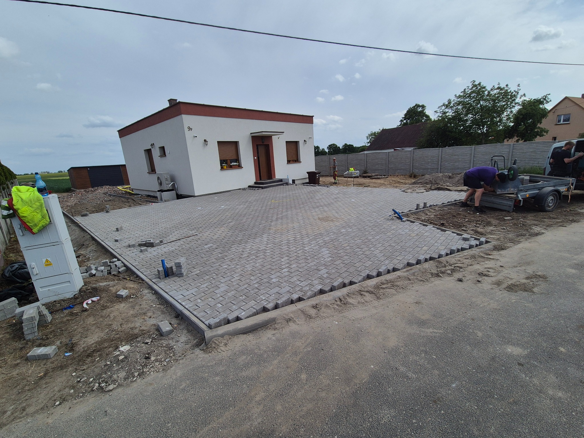 Szara kostka brukowa ułożona na podjeździe przed domem z numerem 9F. W tle widoczny pracownik przygotowujący narzędzia, a obok leżą nieużyte kostki.