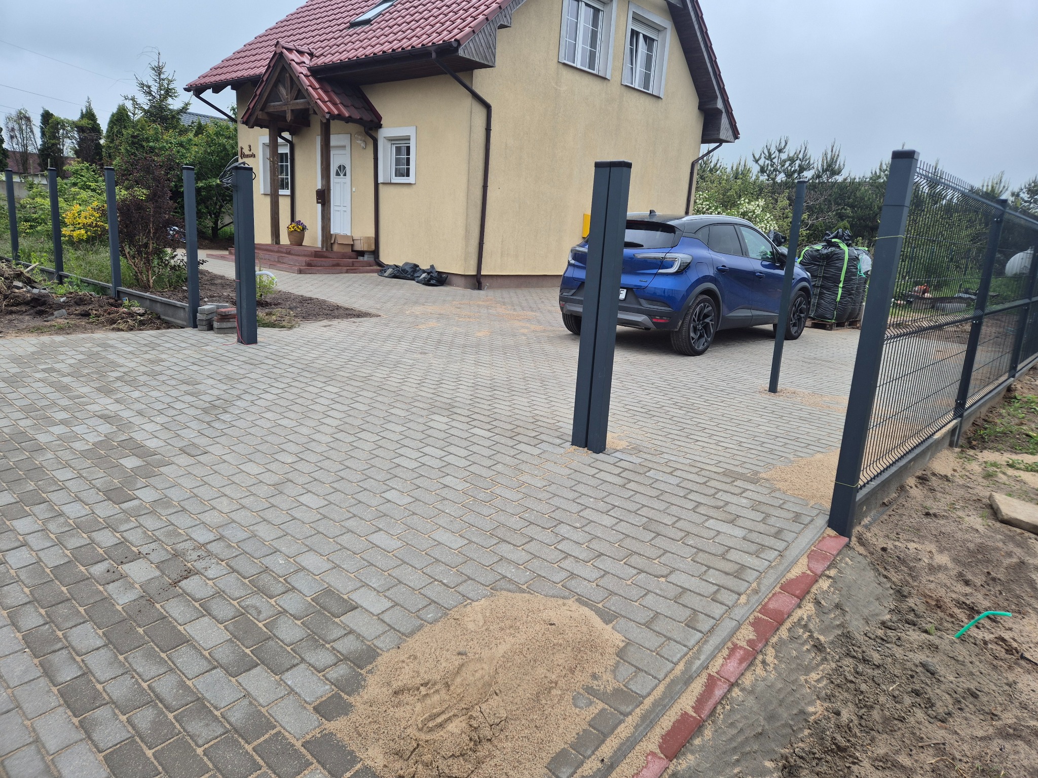 Ułożona kostka brukowa na podjeździe z postawionymi słupkami ogrodzeniowymi i zaparkowanym niebieskim samochodem. Widoczny piasek i obrzeża.