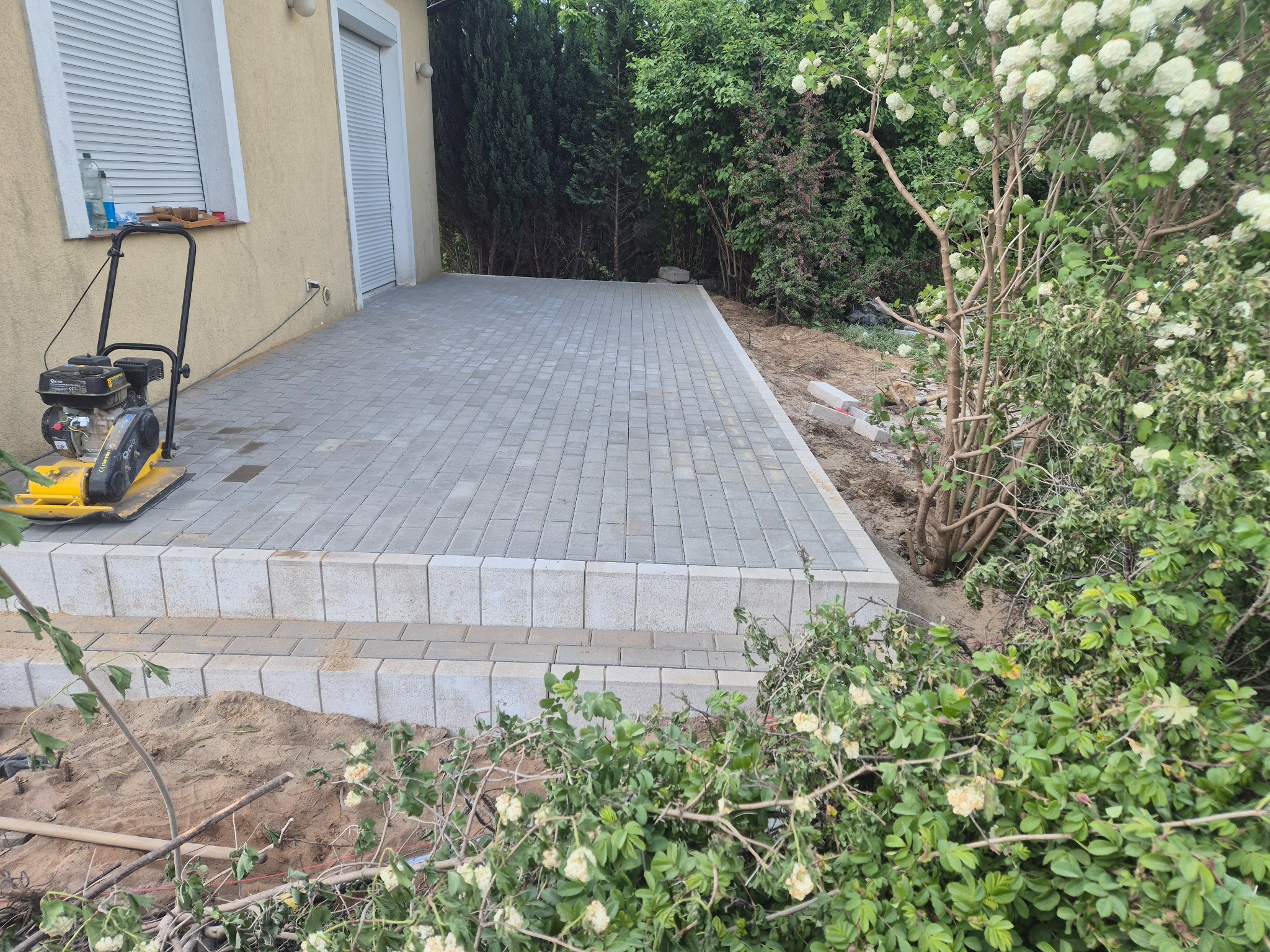 Jasnoszara kostka brukowa ułożona na tarasie przy domu, widoczna zagęszczarka i roślinność. Krawędzie tarasu wykończone kostką. Częściowo widoczny budynek.