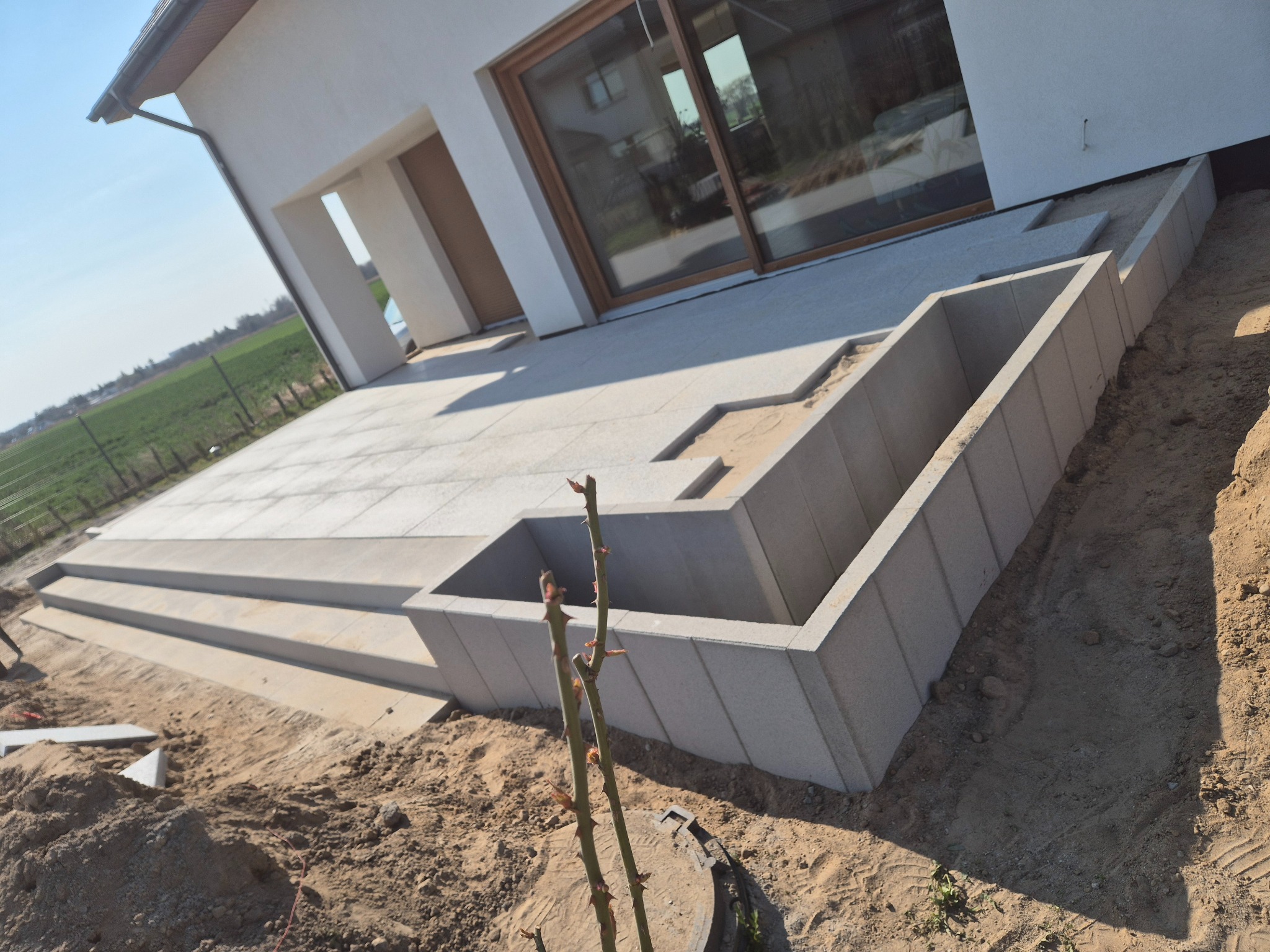Jasnoszare płyty tarasowe przy nowoczesnym domu, z elementami murków i schodów w trakcie budowy, ziemia wokół, widok lekko z boku.