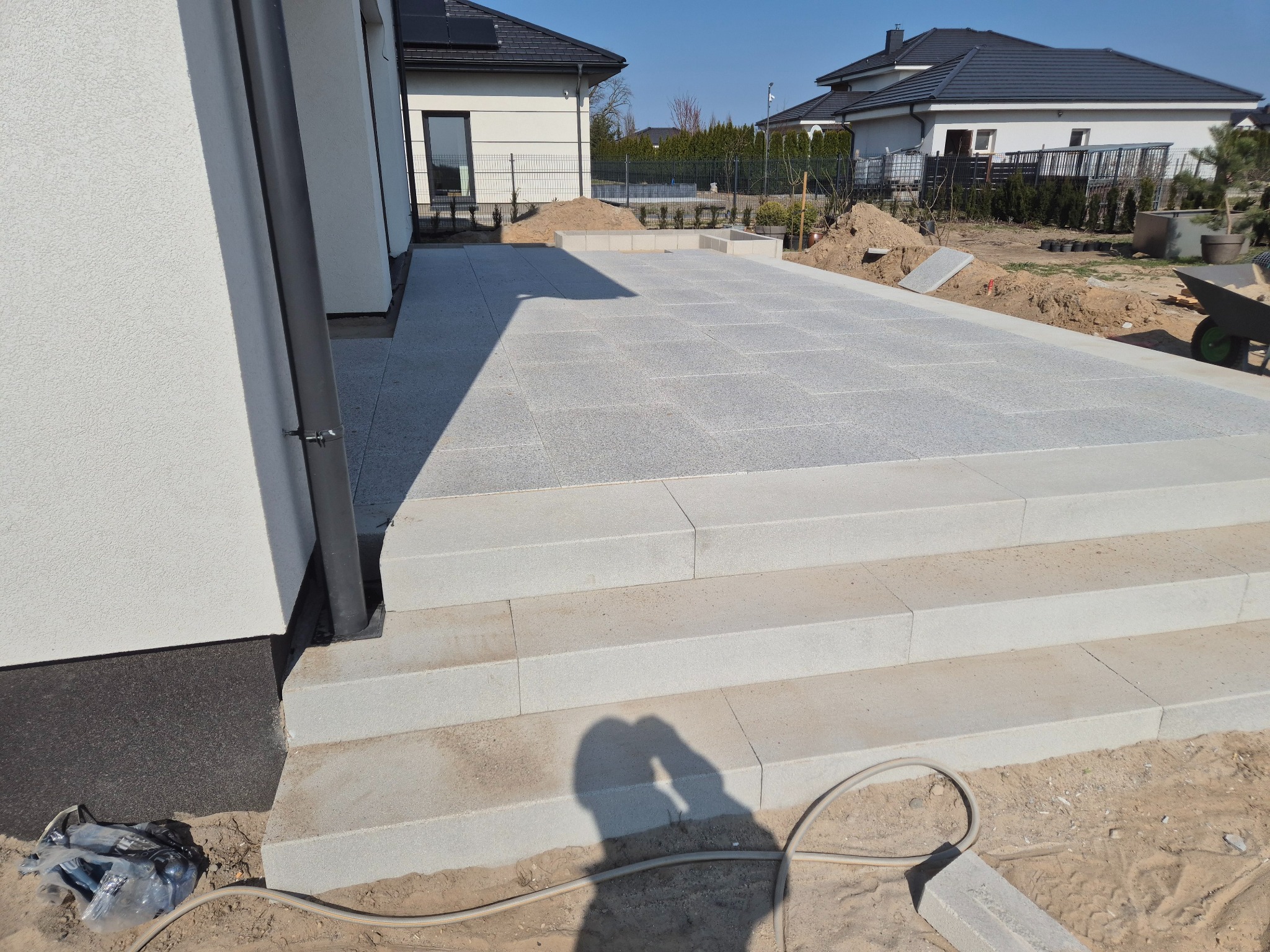 Jasnoszara, ułożona kostka granitowa tworzy taras z trzema schodami. W tle dom z solarnymi panelami i przygotowany teren pod nasadzenia.