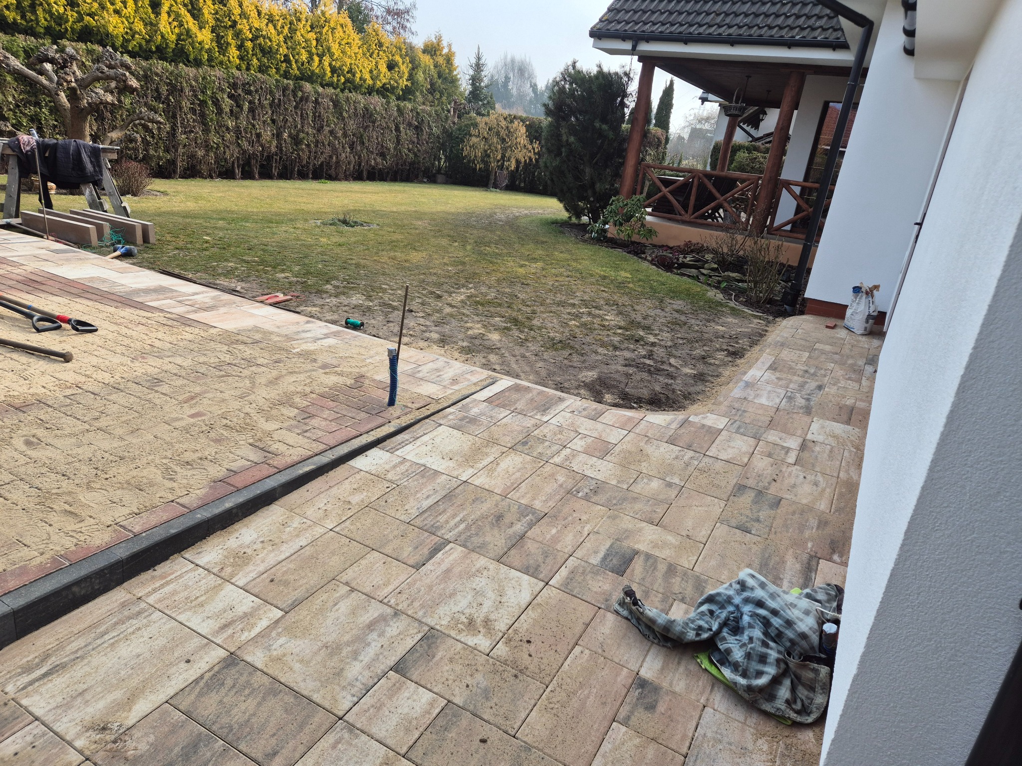 Ułożona kostka brukowa w ogrodzie, z widocznym piaskiem i obrzeżami. Część terenu przygotowana pod dalsze prace. W tle budynek mieszkalny i zieleń.