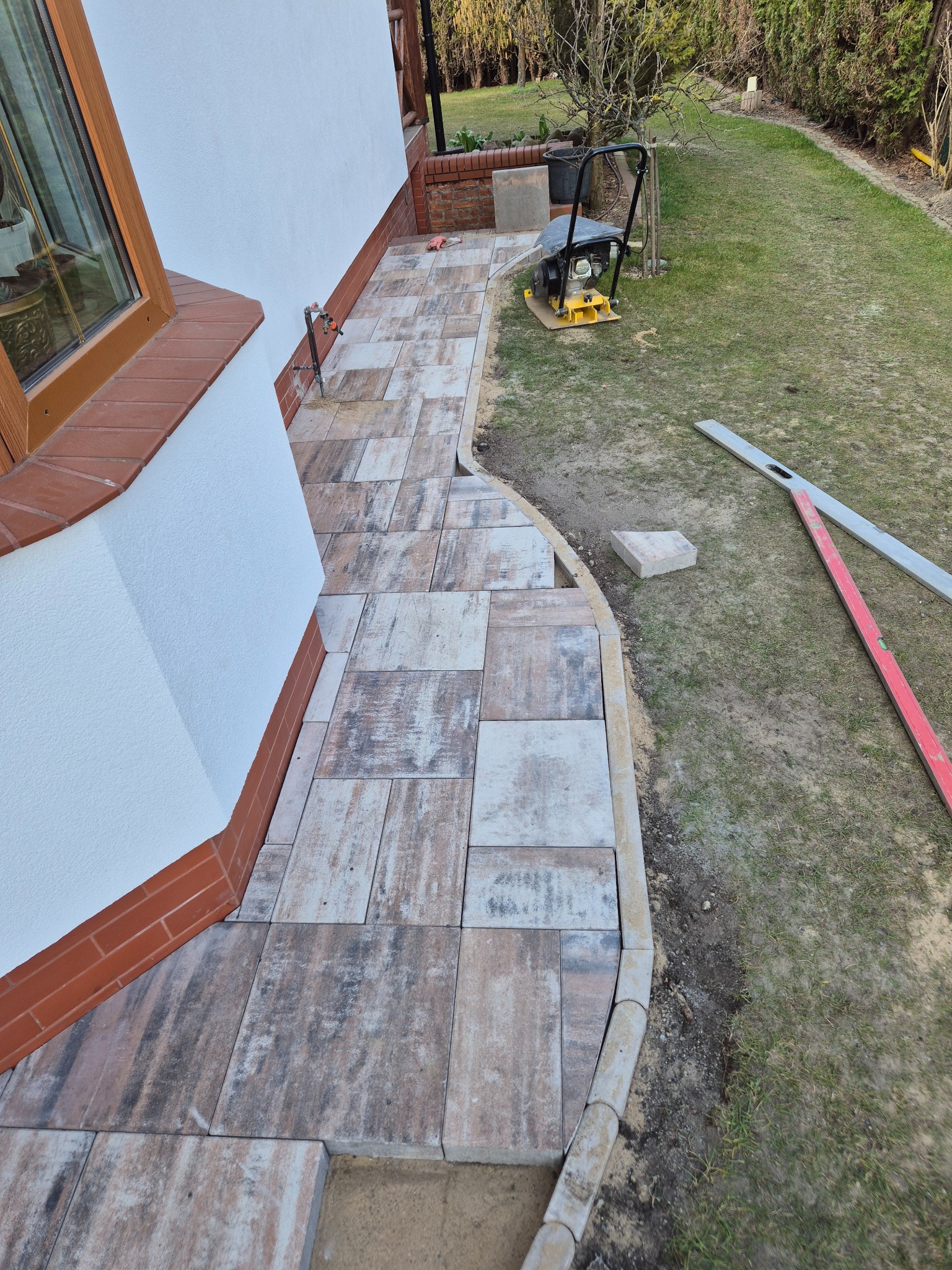 Ułożona ścieżka z kostki brukowej w odcieniach beżu i szarości, obok domu z zielonym trawnikiem i ubijarką w tle. Widoczne narzędzia i krawężnik.
