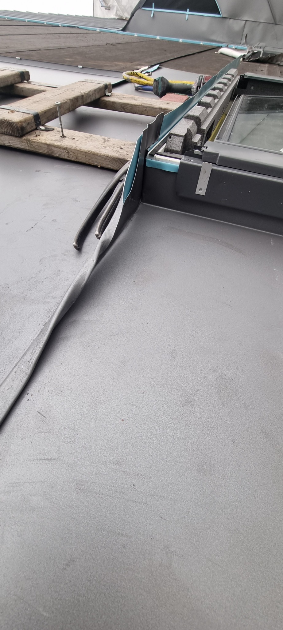 Czarny dach z blachy trapezowej, obróbka blacharska okna dachowego w trakcie montażu, widoczne narzędzia i drewniane podpory.