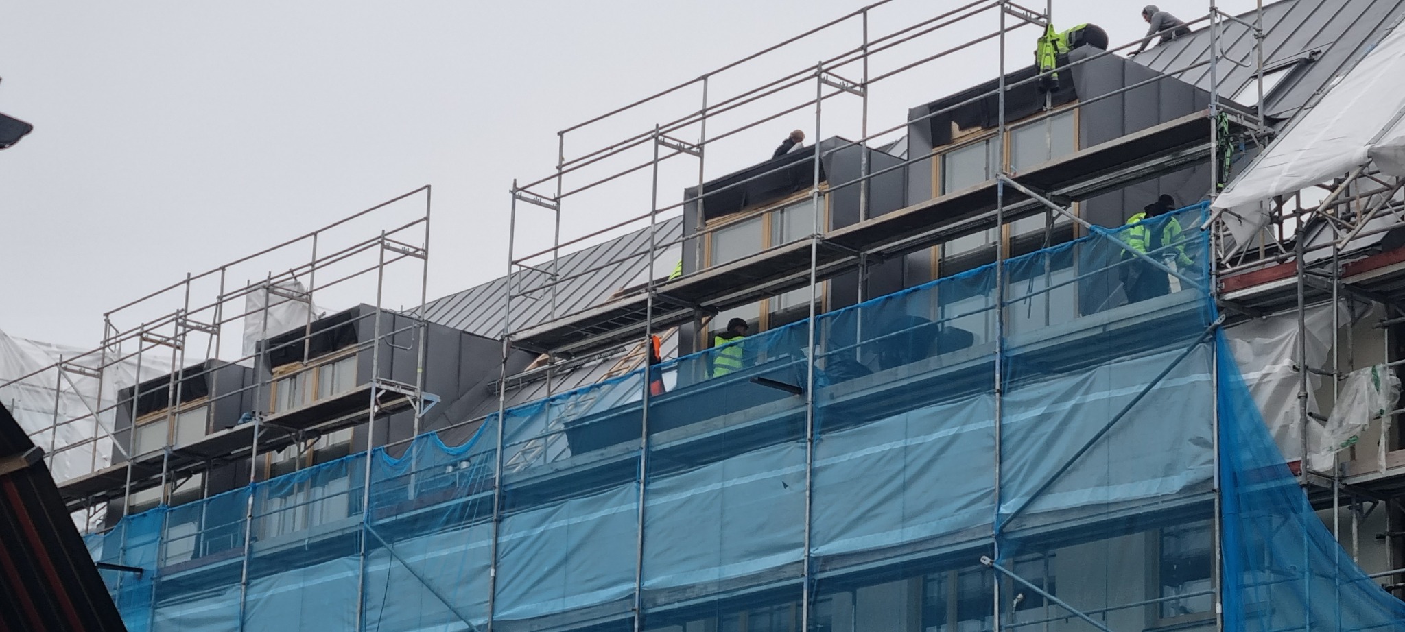 Ekipa dekarzy na rusztowaniu montuje metalowy dach na budynku z dużymi oknami, zabezpieczonym niebieską siatką ochronną.